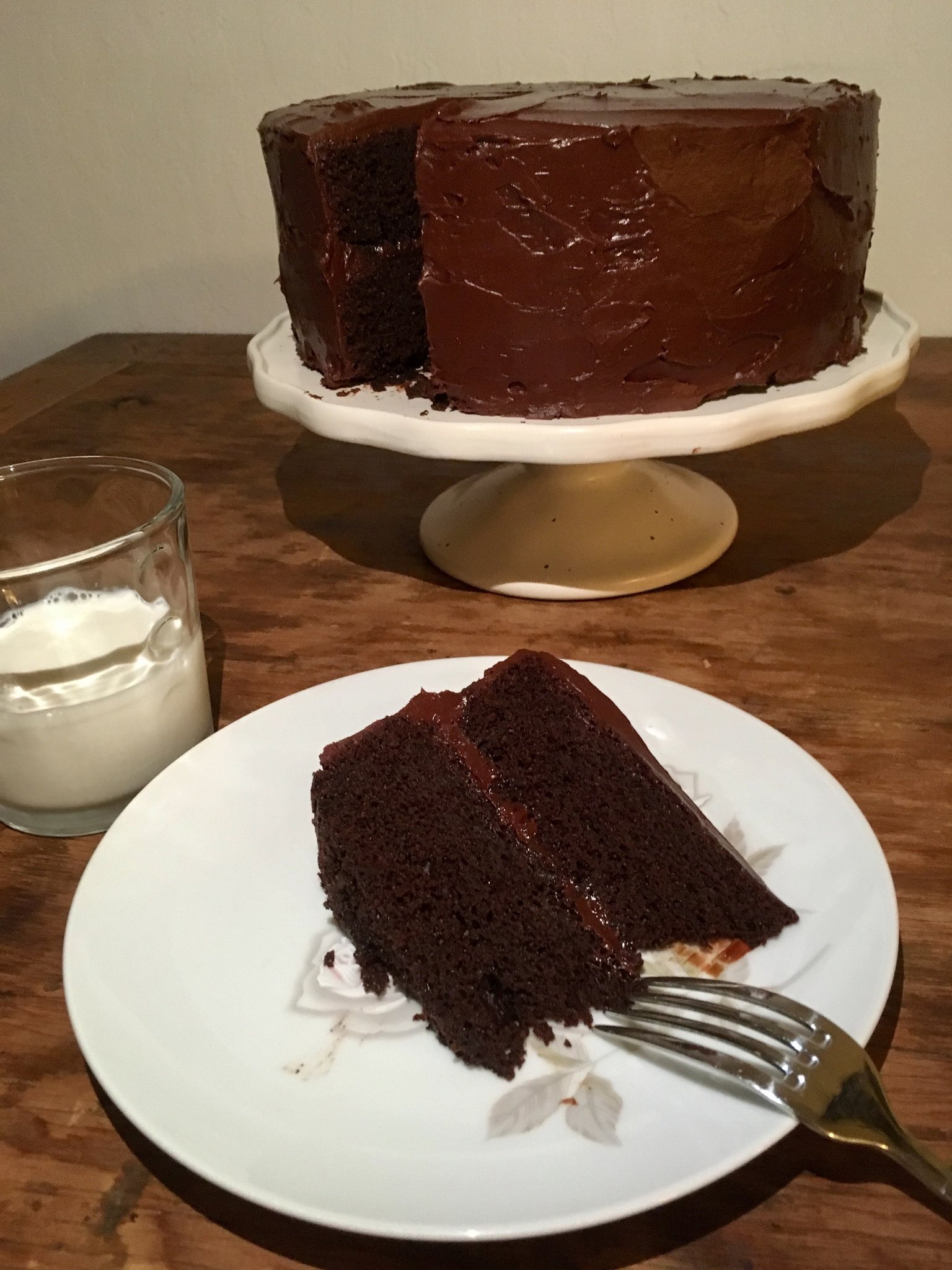 A truly stunning and delicious-looking chocolate cake on a stand, with a slice taken out of it and presented on a plate in front of the cake.